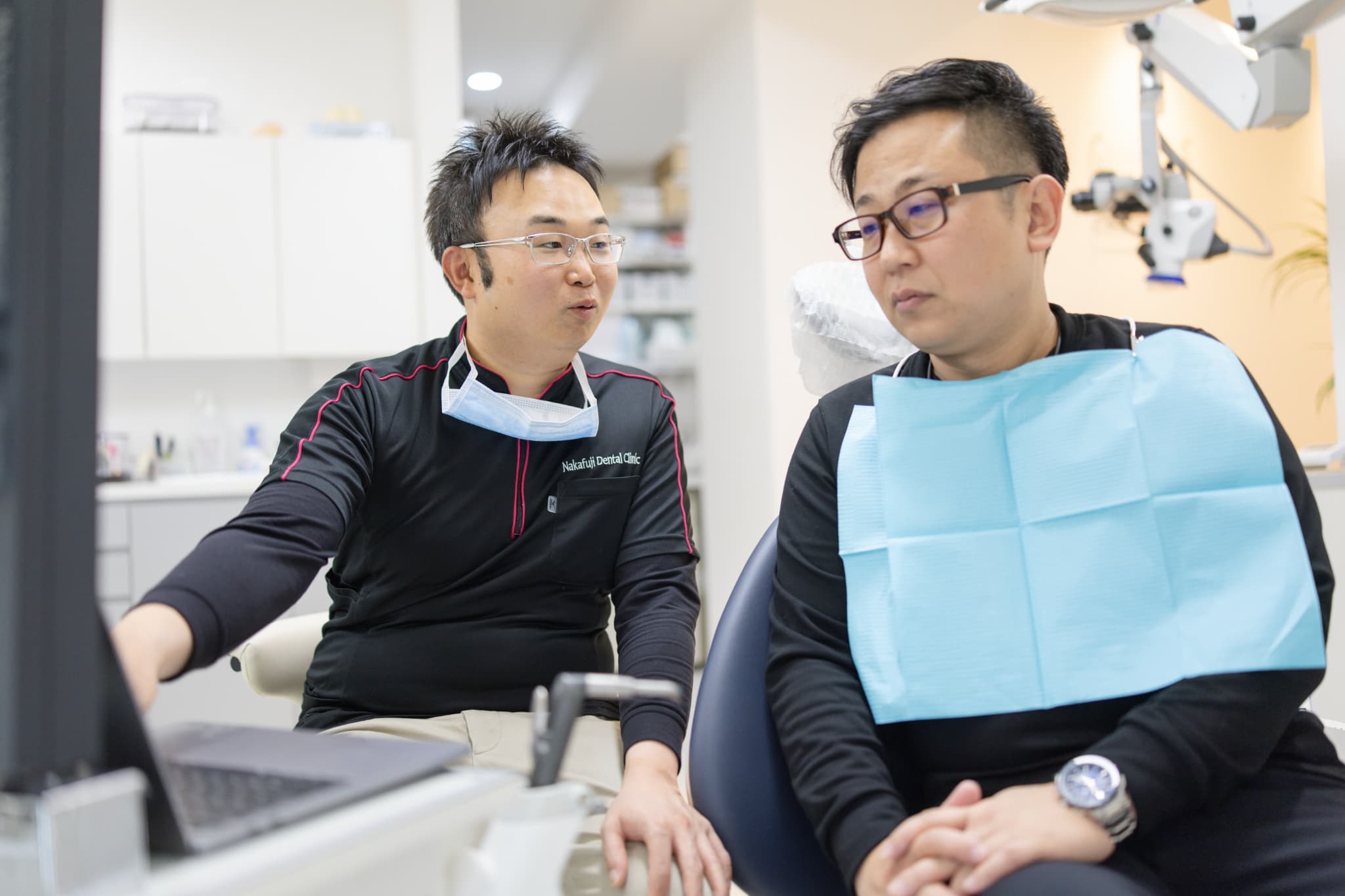 診療チェアで男性患者と一緒にモニターを見ながら治療内容を説明する男性歯科医師のカウンセリング風景