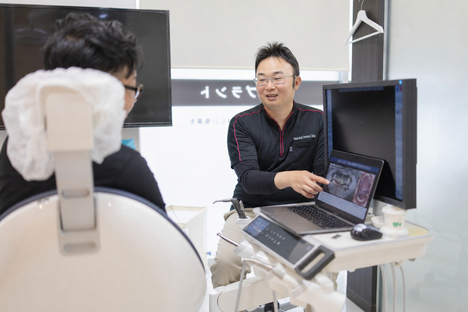 ノートパソコンの歯の画像を見せながら患者に笑顔で治療を説明する男性歯科医師のカウンセリング風景