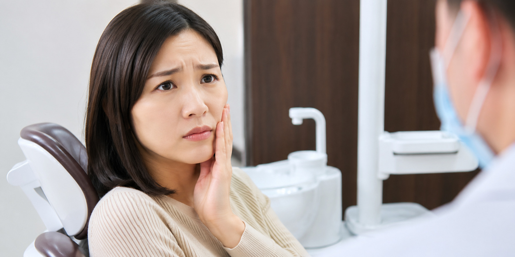 診療チェアで頬に手を当て、歯の痛みを歯科医師に相談するつらそうな表情の女性患者